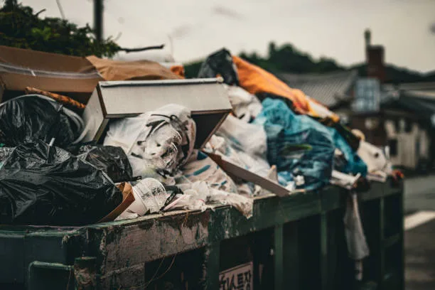 A crowded dumpster brimming with various types of trash, including black garbage bags, cardboard, and plastic waste, situated in an urban area. The image captures a pressing environmental issue of waste management in cities. Highlights the impact of inadequate disposal systems on public cleanliness and sustainability.